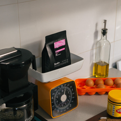 A bag of Bright Eyes sitting in kitchen scales on a counter next to a pot of coffee