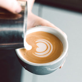 A person pouring steamed milk into a cup of espresso to create a pattern on the surface.