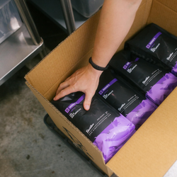 A person's hand reaching into a cardboard box filled bags of Ronnies.